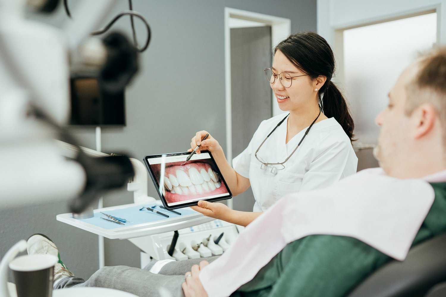 Dr. Yanfei Jiang erklärt einem Patienten im Behandlungsstuhl ein Zahnbild auf dem Tablet